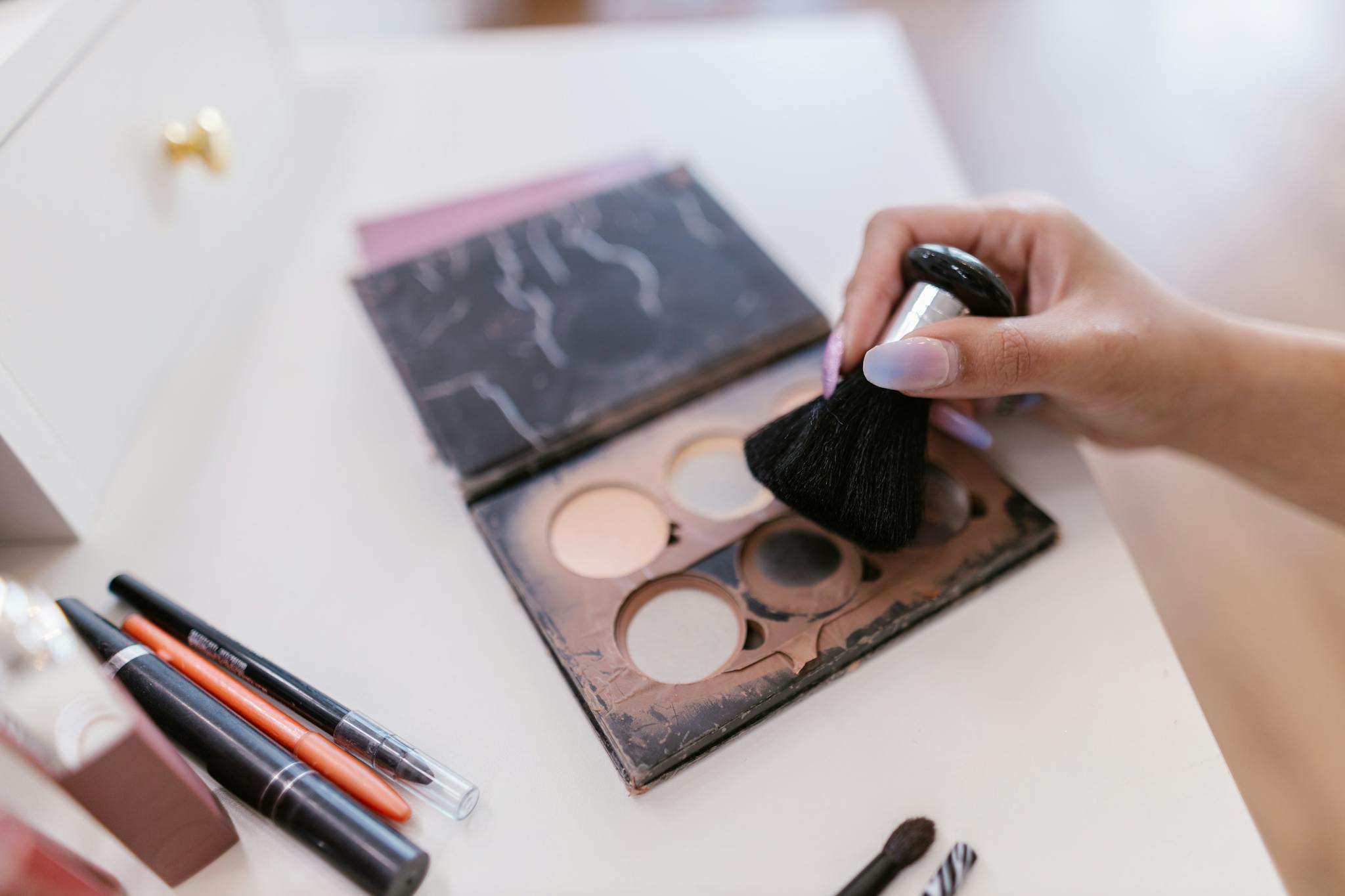 A hand holding a makeup brush over an open palette with brushes nearby on a table.